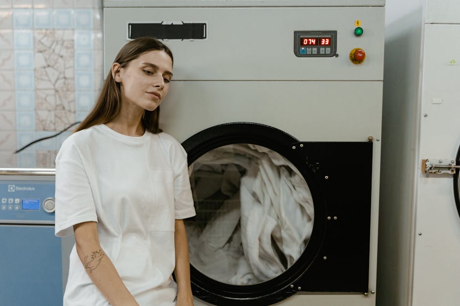 A woman with long brown hair, dressed in a white cleaning uniform, stands beside a commercial industrial washing machine with a glass door, which reveals white laundry inside. She appears to be inspec