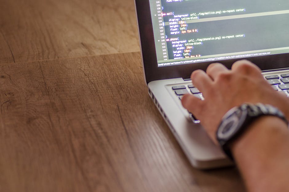 Close-up of a person's hands typing on a MacBook Pro laptop displaying a dark-themed coding interface with lines of colored code. The laptop is placed on a black table in a dimly lit setting, with the