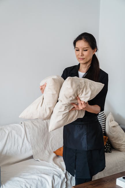 Close-up of a person's hand holding a handheld vacuum cleaner, actively vacuuming a green upholstered armchair in a living room setting. The armchair has a tufted backrest with square stitching and is